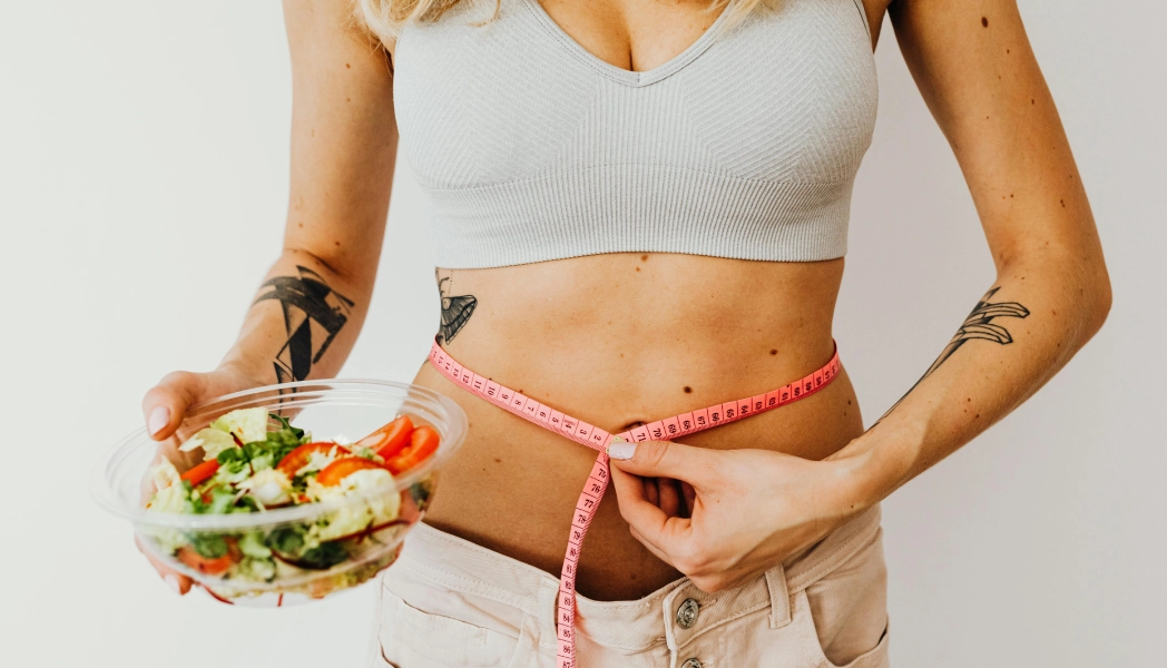 A person measuring their waist with a tape while holding a bowl of salad.