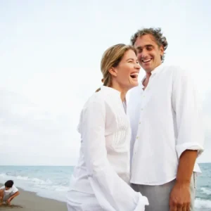 A happy couple laughing on the beach in white clothing.