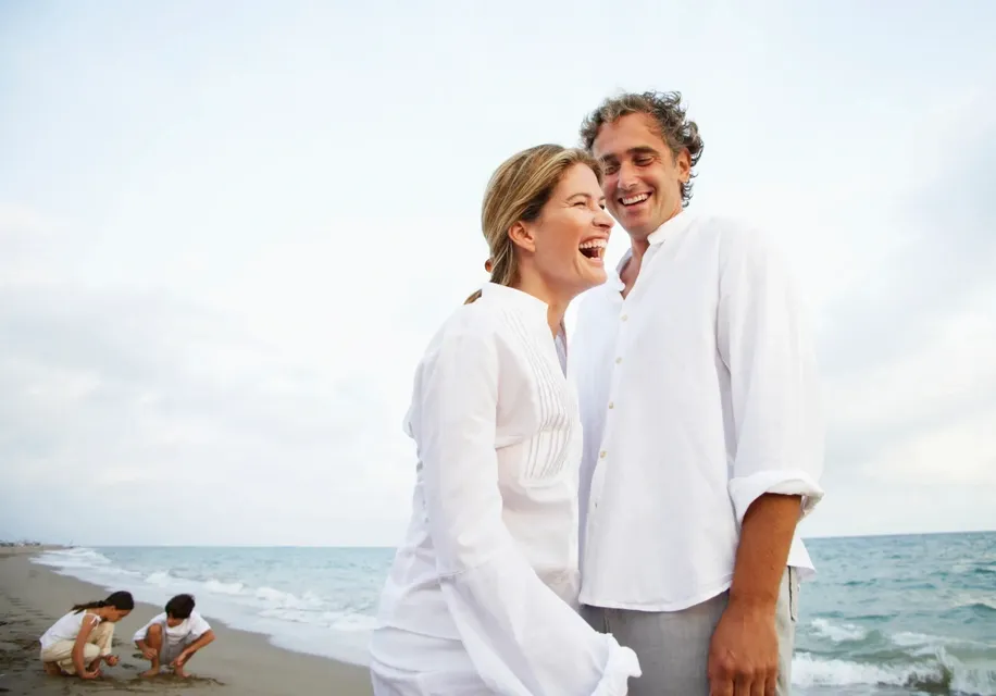 A happy couple laughing on the beach in white clothing.
